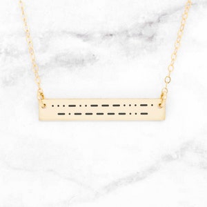 May include: A gold chain necklace with a bar pendant featuring a message in braille.
