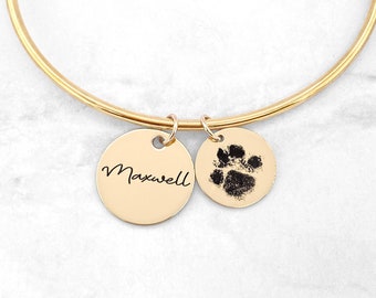 Könnte beinhalten: Goldenes Armreif-Armband mit zwei Anhängern. Ein Anhänger ist mit dem Namen "Maxwell" graviert, der andere Anhänger hat einen schwarzen Pfotenabdruck.