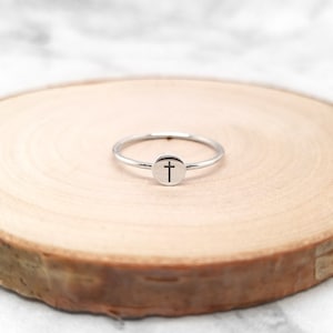 May include: A silver ring with a small circle design featuring a cross. The ring is displayed on a wooden surface.