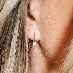 Op de afbeelding: Close-up van een gouden oorring met een kleine, heldere, ronde edelsteen. De oorring wordt gedragen aan een oor, met blond haar op de achtergrond. Het ontwerp is eenvoudig en elegant.