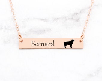 May include: Rose gold bar necklace with the name "Bernard" engraved on it, along with a black silhouette of a dog. The necklace has a delicate chain and is displayed on a white marble surface.