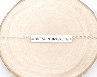 Puede incluir: Un collar de plata con un colgante rectangular que dice "36°9'57" N 86°46'40" W" sobre una superficie de madera.