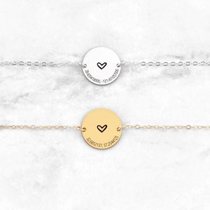 May include: Two necklaces with a circular pendant featuring a small heart and coordinates engraved on the front. The top necklace has a silver chain and pendant, while the bottom necklace has a gold chain and pendant.