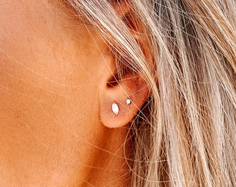 May include: Close-up of a person's ear with two small silver stud earrings. The earrings are set with clear stones.