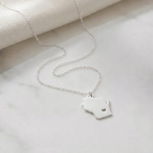 May include: A silver necklace featuring a pendant shaped like the state of Wisconsin with a heart cutout. The chain and pendant are made of a shiny, reflective metal. The necklace is displayed on a white surface.