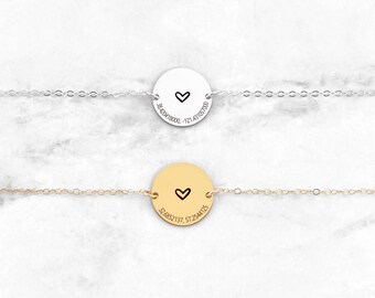 May include: Two necklaces with a circular pendant featuring a small heart and coordinates engraved on the front. The top necklace has a silver chain and pendant, while the bottom necklace has a gold chain and pendant.