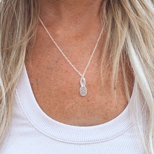 May include: A silver chain necklace with a pineapple pendant. The necklace is worn by a person with long blonde hair.