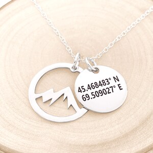 May include: A silver necklace with two pendants. One pendant is a circle with a mountain cutout, and the other is a round tag with coordinates: 45.468483&deg; N 69.509027&deg; E. The necklace is on a light wood surface.