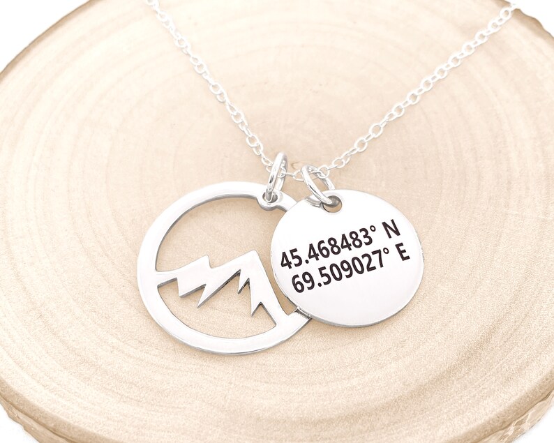 May include: A silver necklace with two pendants. One pendant is a circle with a mountain cutout, and the other is a round tag with coordinates: 45.468483&deg; N 69.509027&deg; E. The necklace is on a light wood surface.