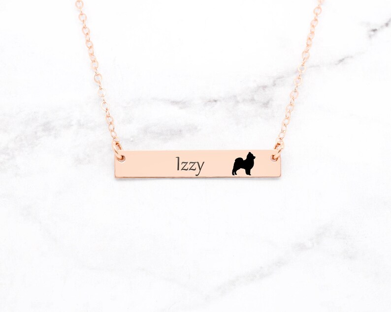 May include: A rose gold bar necklace with the name "Izzy" engraved on it, along with a black silhouette of a dog. The necklace is displayed on a white and gray marble surface.