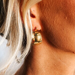 May include: Close-up of a gold-colored, chunky hoop earring. The earring is a thick, rounded shape with a polished finish. The earring is worn on the ear of a person with blonde hair.