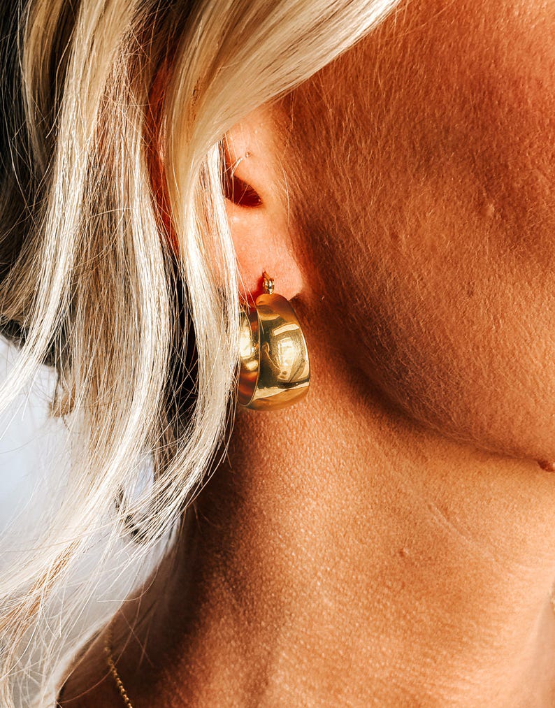May include: Close-up of a gold-colored, chunky hoop earring. The earring is a thick, rounded shape with a polished finish. The earring is worn on the ear of a person with blonde hair.