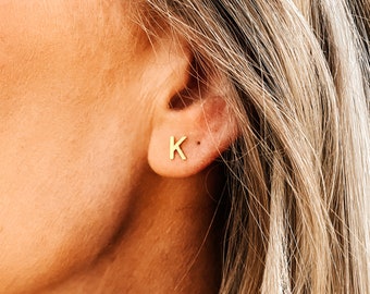 Puede incluir: Primer plano de una oreja con un pendiente de botón con la letra "K" dorada. El pendiente es pequeño y sencillo, con un diseño minimalista. La piel de la persona es clara y su pelo es rubio.