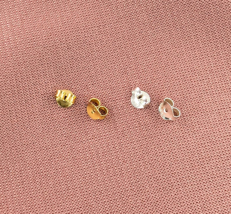 May include: Four earring backs, two gold and two silver, on a pink fabric background.