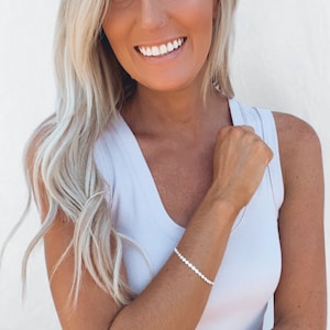 Op de afbeelding: Een vrouw met lang blond haar draagt een wit tanktop en een zilveren kralen armband. Ze glimlacht en kijkt naar de camera.