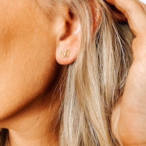 May include: A close-up of a person's ear with a small gold butterfly stud earring. The earring is delicate and feminine.