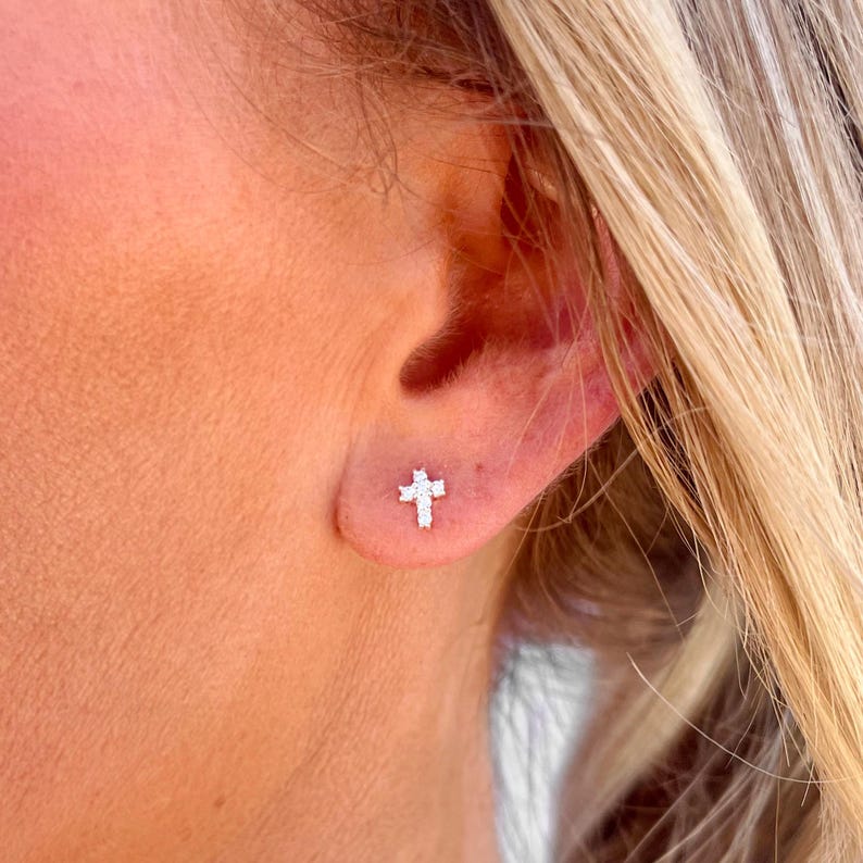 May include: A close-up of a person's ear with a small, silver cross earring set with tiny, clear cubic zirconia stones.