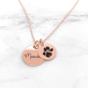 May include: Rose gold necklace with two charms. One charm is a round disc with the name "Maxwell" engraved on it. The other charm is a round disc with a paw print.