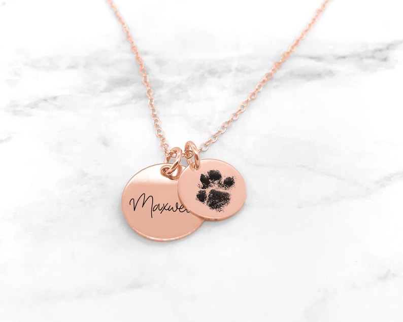 May include: Rose gold necklace with two charms. One charm is a round disc with the name "Maxwell" engraved on it. The other charm is a round disc with a paw print.