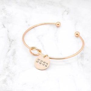 Op de afbeelding: Een gouden armband met een knoopdetail en een ronde bedel met gegraveerde coördinaten. De bedel geeft "15° 23' 45" N 23° 56' 13" W" aan.