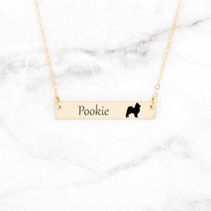 May include: A gold-colored bar necklace with the name "Pookie" engraved on it, accompanied by a black silhouette of a dog. The necklace hangs from a delicate gold chain, set against a white marble background.