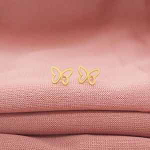 May include: A pair of gold butterfly-shaped earrings on a pink fabric background.