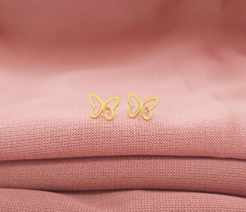 May include: A pair of gold butterfly-shaped earrings on a pink fabric background.