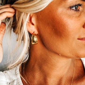 May include: Gold-toned, chunky hoop earrings. The earrings have a squared-off shape and a polished finish. A delicate gold necklace is also visible. The image shows a close-up of a person's face and neck, with blonde hair.