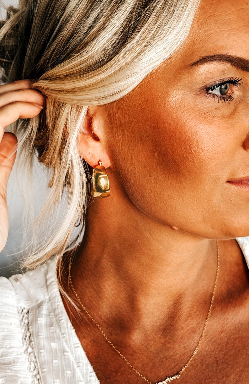 May include: Gold-toned, chunky hoop earrings. The earrings have a squared-off shape and a polished finish. A delicate gold necklace is also visible. The image shows a close-up of a person's face and neck, with blonde hair.