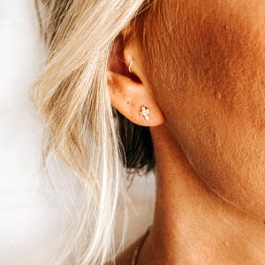 May include: A close-up of a person's ear with a small gold cross earring. The earring has a single clear stone in the center.