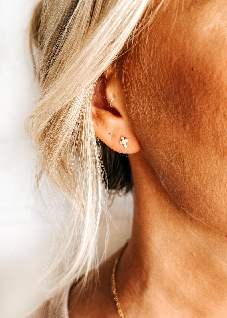 May include: A close-up of a person's ear with a small gold cross earring. The earring has a single clear stone in the center.