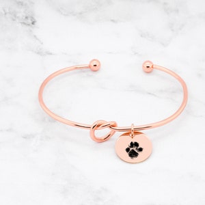 May include: Rose gold bangle bracelet with a knot design and a charm featuring a paw print.