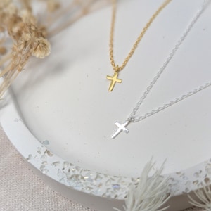 K&ouml;nnte beinhalten: Zwei zarte Halsketten mit Kreuzanh&auml;ngern. Eine Kette ist goldfarben, die andere silberfarben. Beide Halsketten werden auf einer wei&szlig;en, strukturierten Oberfl&auml;che mit getrockneten floralen Akzenten im Hintergrund pr&auml;sentiert. Die Kreuzanh&auml;nger sind klein und schlicht gestaltet.