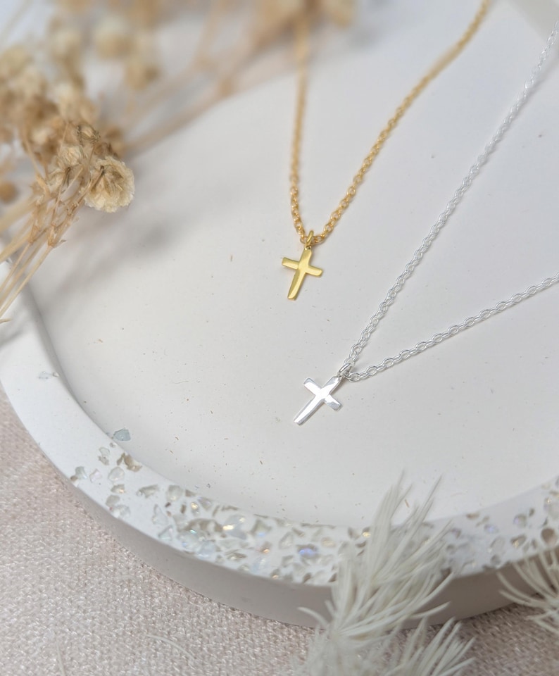 K&ouml;nnte beinhalten: Zwei zarte Halsketten mit Kreuzanh&auml;ngern. Eine Kette ist goldfarben, die andere silberfarben. Beide Halsketten werden auf einer wei&szlig;en, strukturierten Oberfl&auml;che mit getrockneten floralen Akzenten im Hintergrund pr&auml;sentiert. Die Kreuzanh&auml;nger sind klein und schlicht gestaltet.