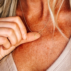 K&ouml;nnte beinhalten: Eine zarte goldfarbene Halskette mit einem kleinen Kreuzanh&auml;nger. Die Halskette wird vor einem Hautton-Hintergrund gezeigt, der die feinen Details des Schmucks hervorhebt. Der Kreuzanh&auml;nger ist ein schlichtes, elegantes Design.
