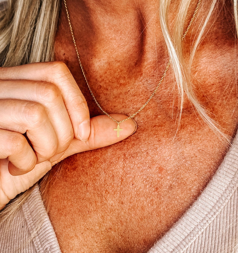 K&ouml;nnte beinhalten: Eine zarte goldfarbene Halskette mit einem kleinen Kreuzanh&auml;nger. Die Halskette wird vor einem Hautton-Hintergrund gezeigt, der die feinen Details des Schmucks hervorhebt. Der Kreuzanh&auml;nger ist ein schlichtes, elegantes Design.