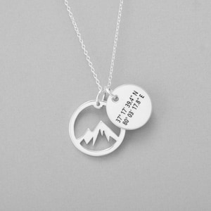 May include: A silver necklace with a mountain range pendant and a round charm. The round charm has the coordinates 37&deg; 17' 39.4" N 80&deg; 03' 17.8" E. The necklace is on a gray background.