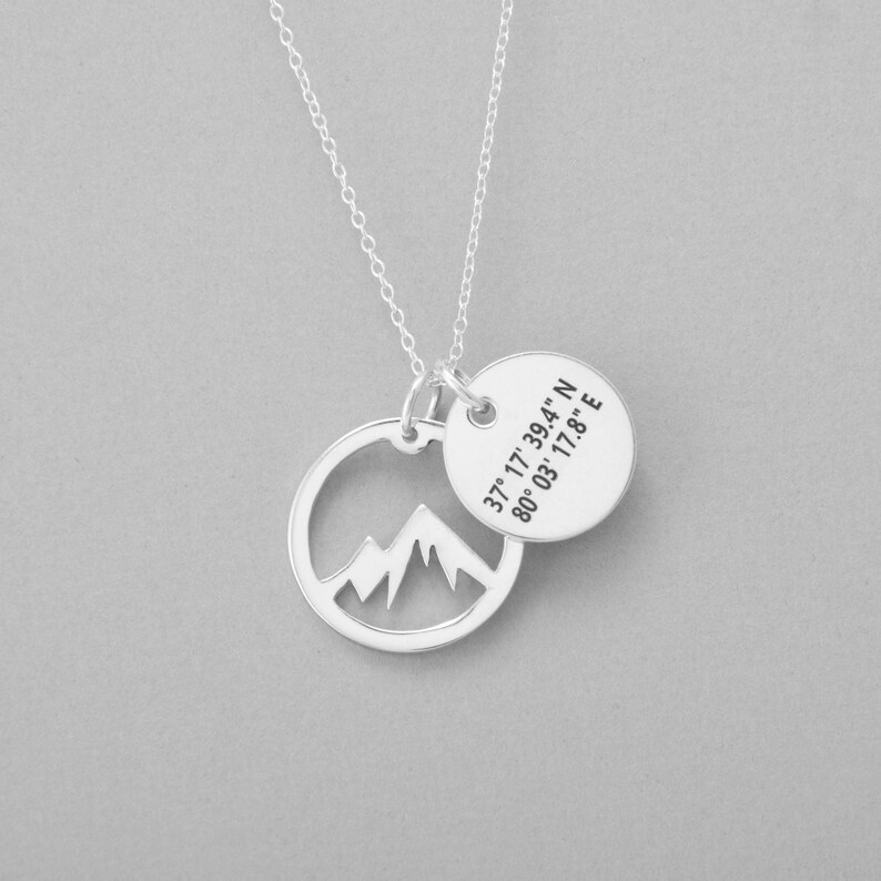 May include: A silver necklace with a mountain range pendant and a round charm. The round charm has the coordinates 37&deg; 17' 39.4" N 80&deg; 03' 17.8" E. The necklace is on a gray background.