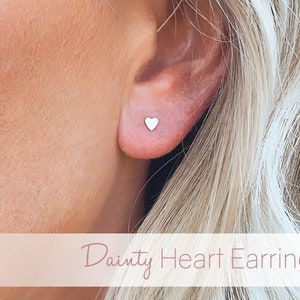 May include: A close-up of a person's ear wearing a pair of dainty silver heart stud earrings.