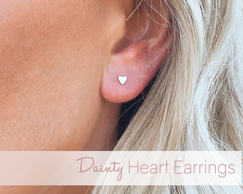 May include: A close-up of a person's ear wearing a pair of dainty silver heart stud earrings.