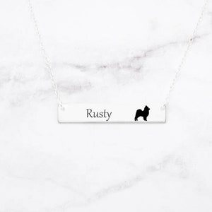 May include: A silver bar necklace with the name "Rusty" engraved on it, accompanied by a black silhouette of a dog. The necklace hangs on a delicate silver chain, set against a white marble background.