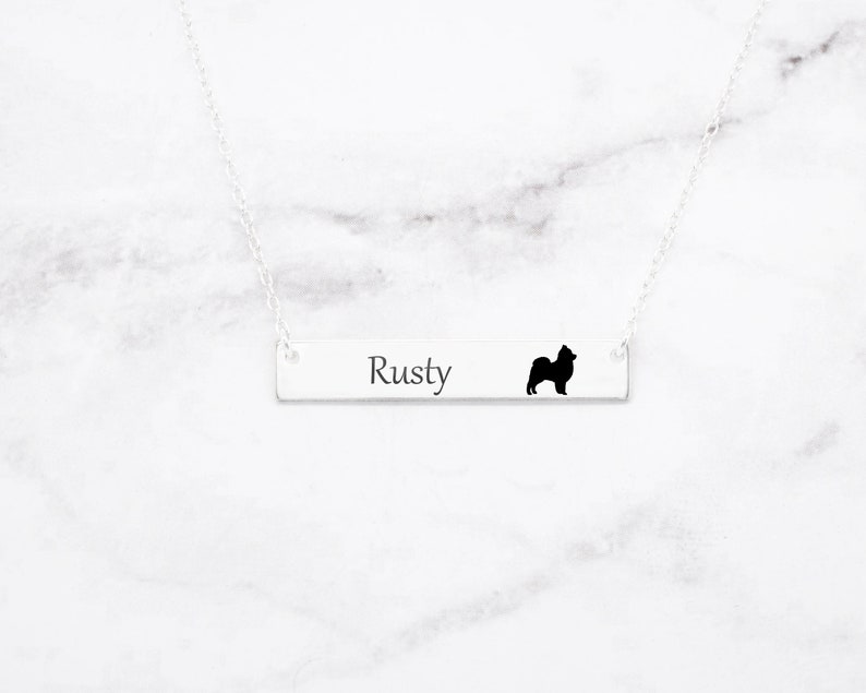 May include: A silver bar necklace with the name "Rusty" engraved on it, accompanied by a black silhouette of a dog. The necklace hangs on a delicate silver chain, set against a white marble background.