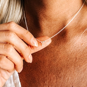 Puede incluir: Un delicado collar de plata con un pequeño colgante con la letra "E" en cursiva. El collar se muestra sobre la piel de una persona, mostrando la fina cadena y el elegante diseño del colgante. El fondo está borroso.