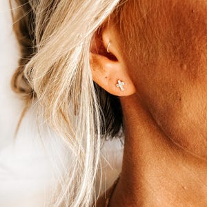 May include: A close-up of a person's ear with a small, diamond-shaped cross earring. The earring is made of gold and has a simple design.