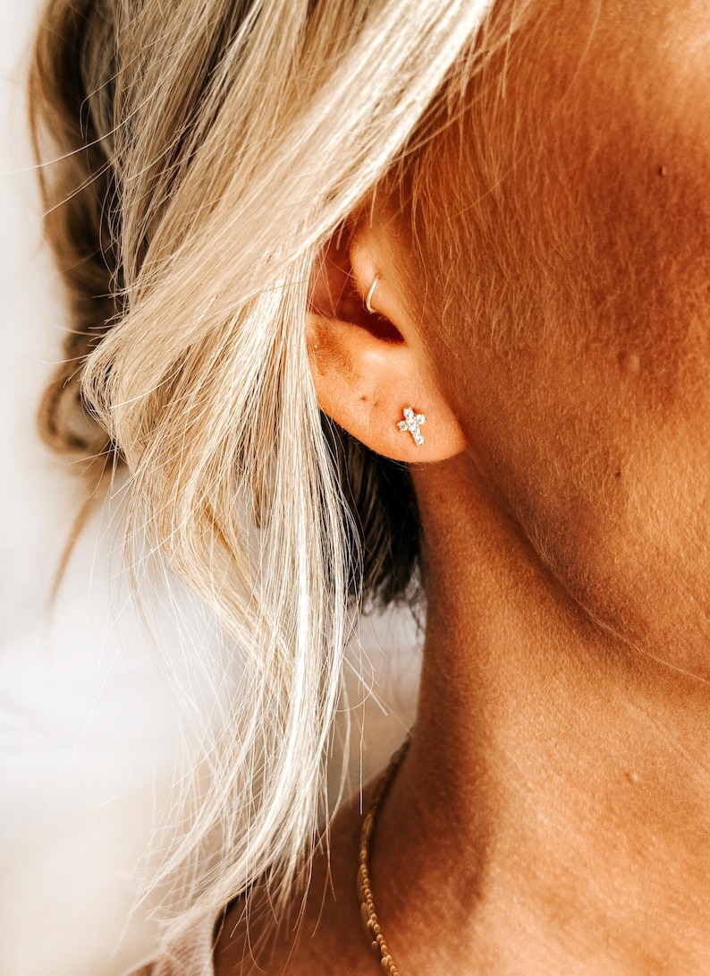 May include: A close-up of a person's ear with a small, diamond-shaped cross earring. The earring is made of gold and has a simple design.