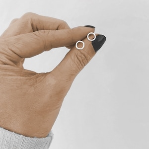May include: A pair of small, silver hoop earrings. The earrings are held in a hand with black nail polish.