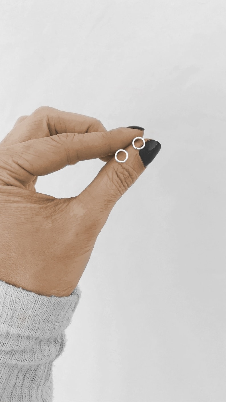 May include: A pair of small, silver hoop earrings. The earrings are held in a hand with black nail polish.