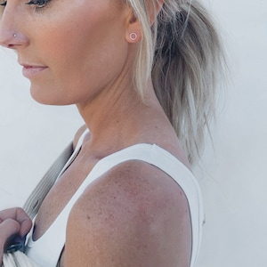 May include: A woman with blonde hair wearing a white vest top and a beige cardigan. She is wearing a small, silver hoop earring in her left ear.