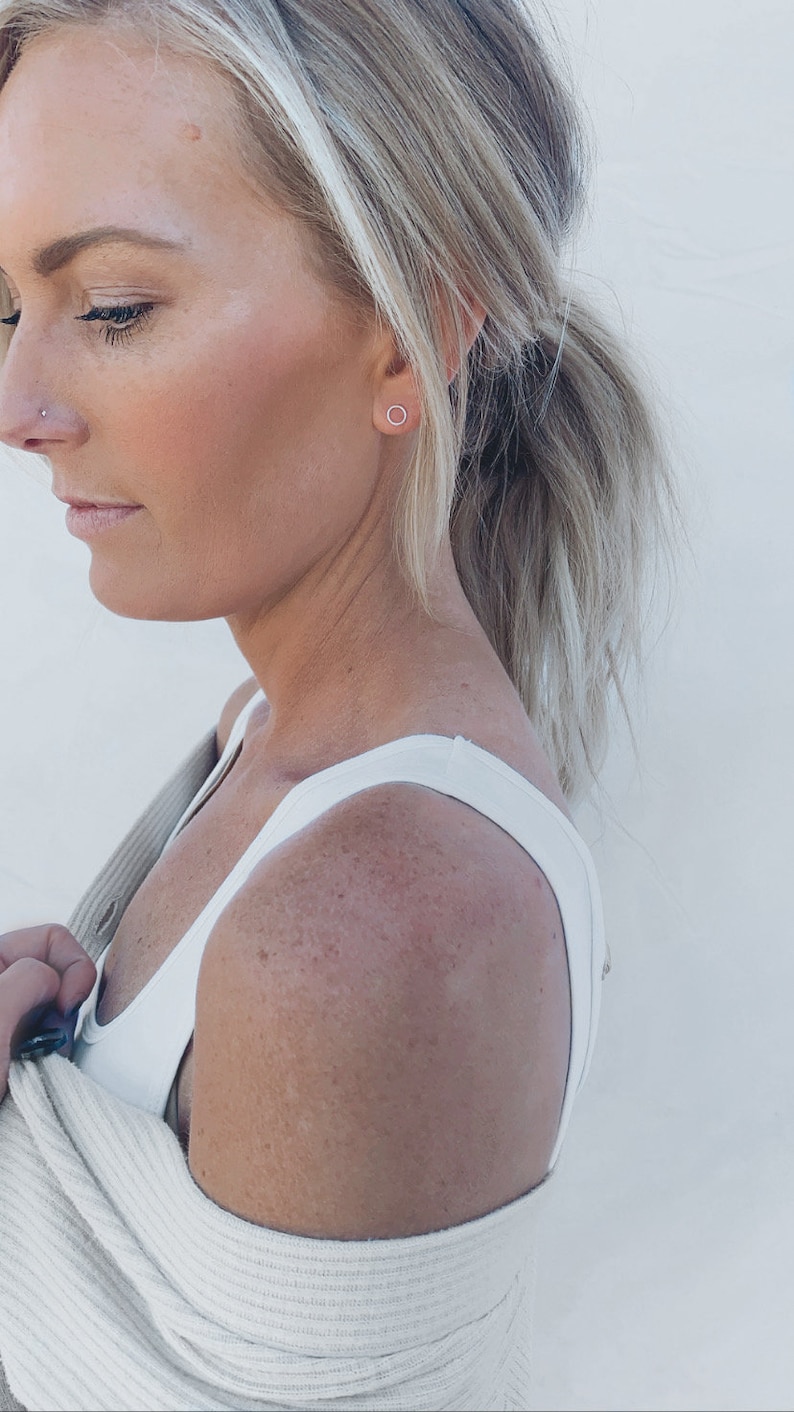 May include: A woman with blonde hair wearing a white vest top and a beige cardigan. She is wearing a small, silver hoop earring in her left ear.