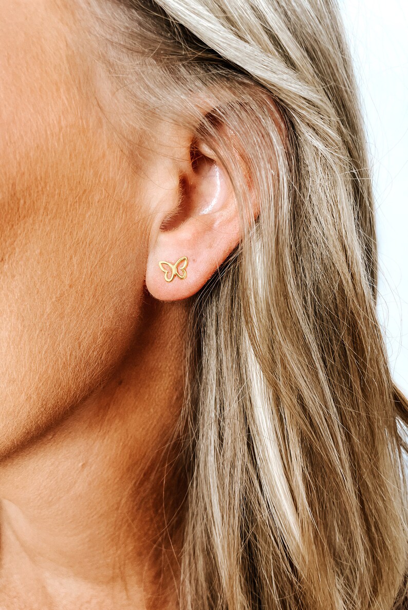 May include: A close-up of a person's ear with a small gold butterfly earring. The earring is a delicate, simple design.